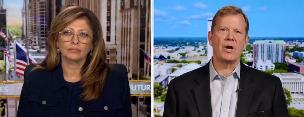 News segment featuring two speakers discussing current events, with a cityscape backdrop and flags in the background.