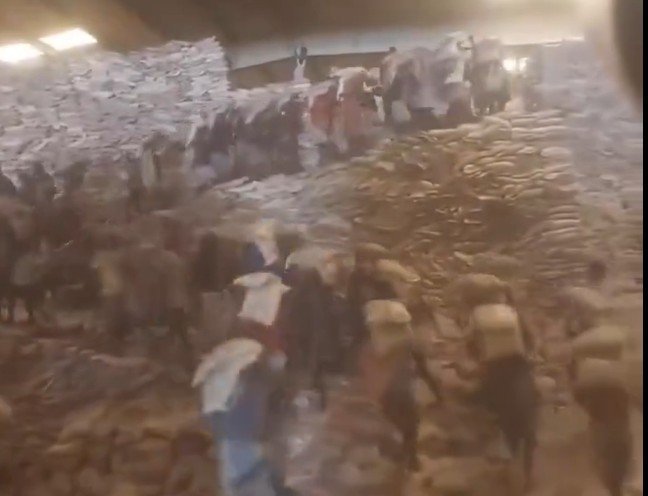 Workers transporting bags of goods in a warehouse with stacked sacks in the background, illustrating labor and logistics in a commercial environment.