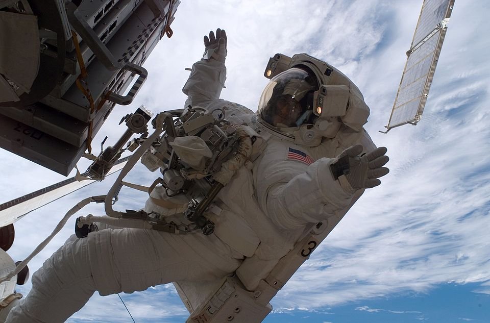 Astronaut in a spacesuit performs a spacewalk outside the International Space Station against a backdrop of Earth and clouds.