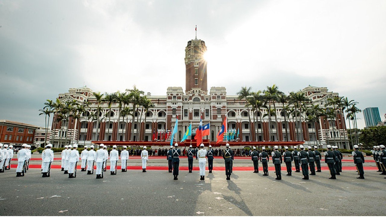 Ceremonial troops in uniform stand in formation in front of a historic government building adorned with flags, set against a cloudy sky.