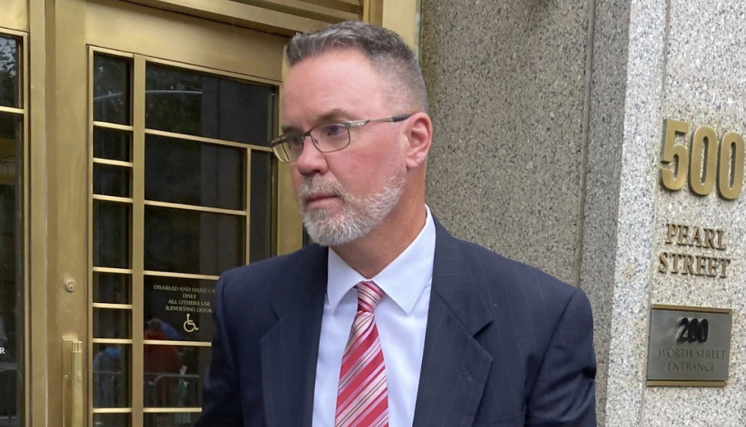 Man in a suit with glasses exits a building at Pearl Street, showcasing a serious expression during a public appearance.