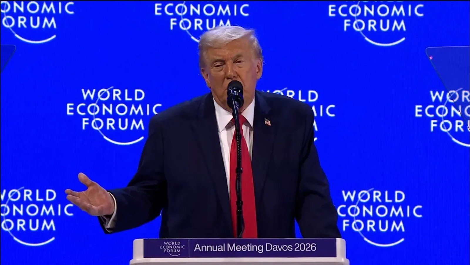 Donald Trump speaking at the World Economic Forum Annual Meeting in Davos 2026, addressing global economic issues with a microphone in hand against a blue backdrop.