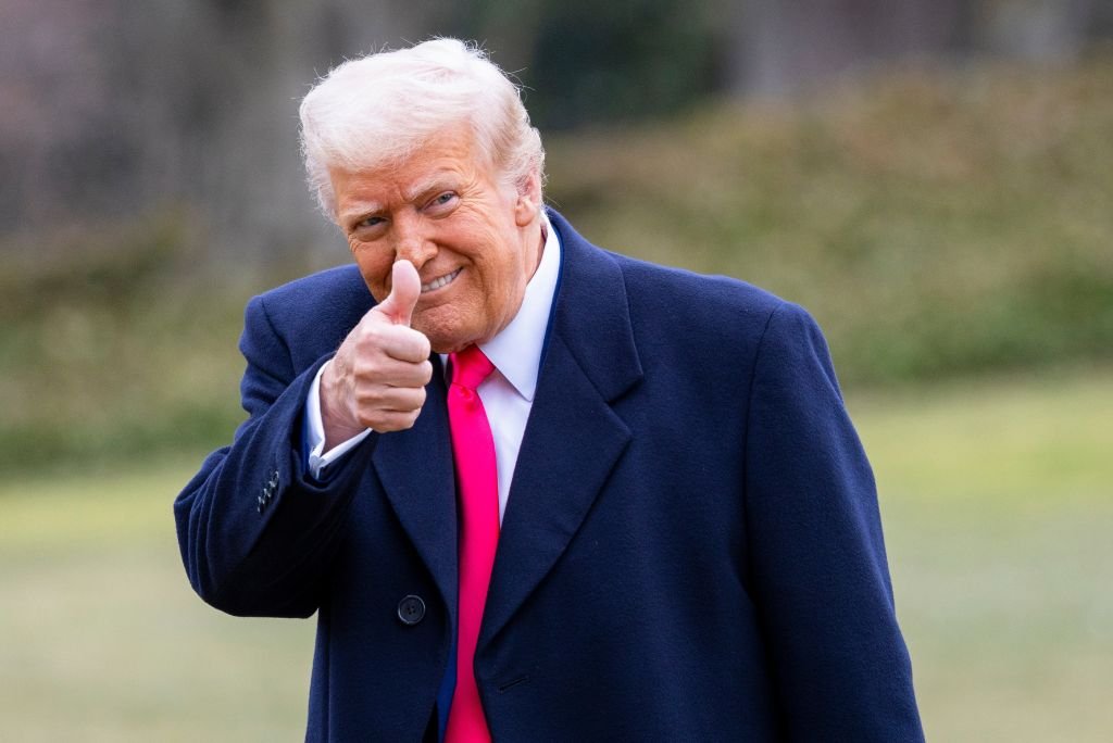 Former President Donald Trump giving a thumbs-up gesture while wearing a navy coat and pink tie outdoors.
