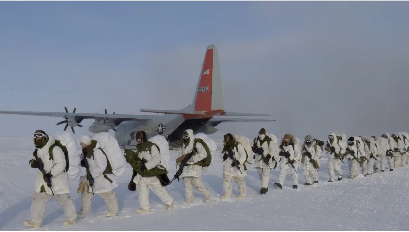 Soldiers in winter gear march across a snowy landscape near a military aircraft, preparing for a cold-weather training exercise.