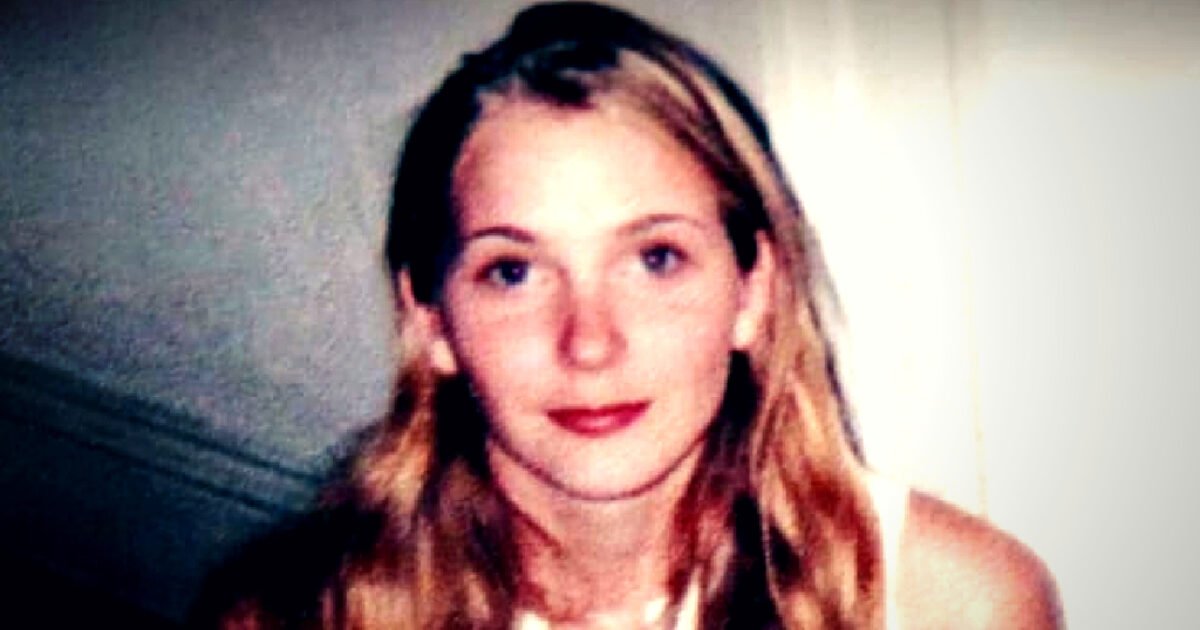 Young woman with long, wavy blonde hair and a neutral expression, posing in a softly lit indoor setting.