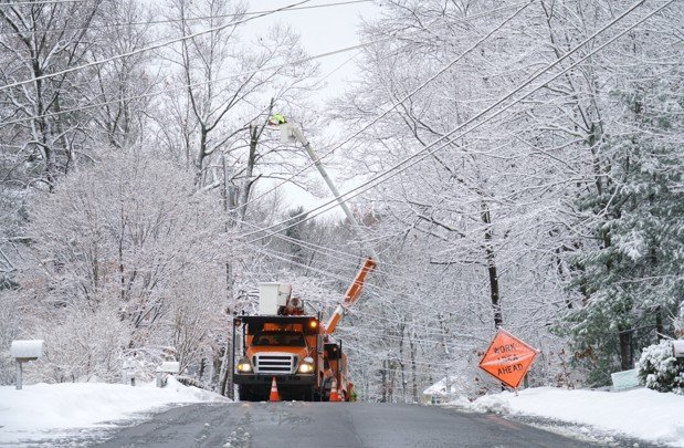Utility workers using a cherry picker to trim trees near power lines on a snowy road, with a caution sign indicating work ahead.