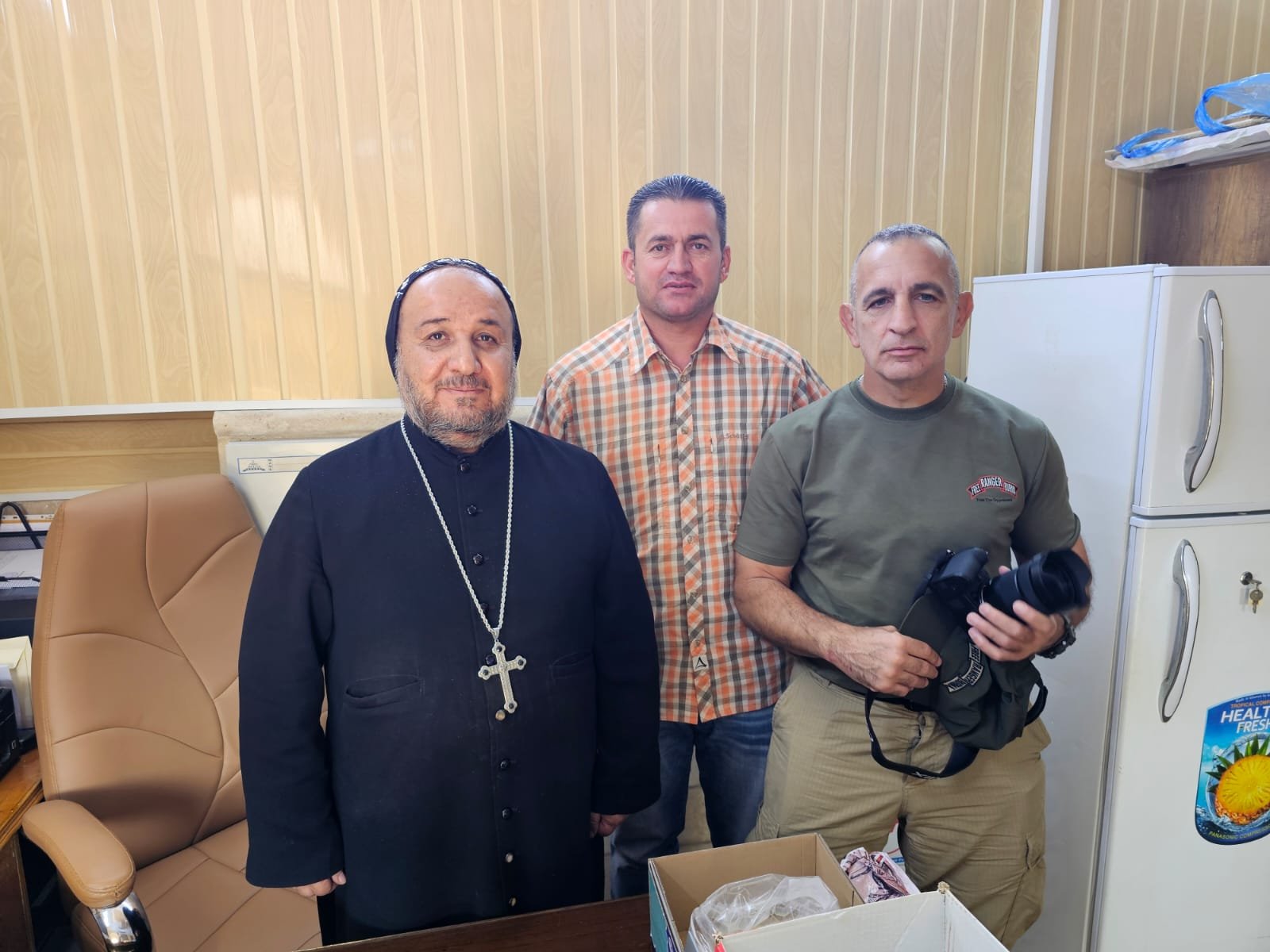 Three men pose together in an office setting, with one wearing religious attire and others holding a camera, highlighting a moment of collaboration.