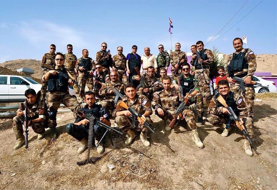 Group of armed soldiers posing for a photo outdoors, showcasing military gear and weapons against a backdrop of hills and a clear sky.