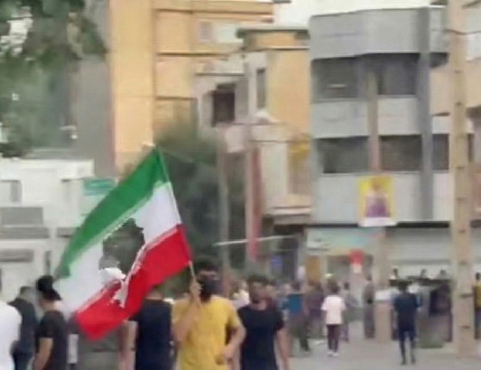 Protesters gather in the street holding an Iranian flag during a demonstration in an urban area.