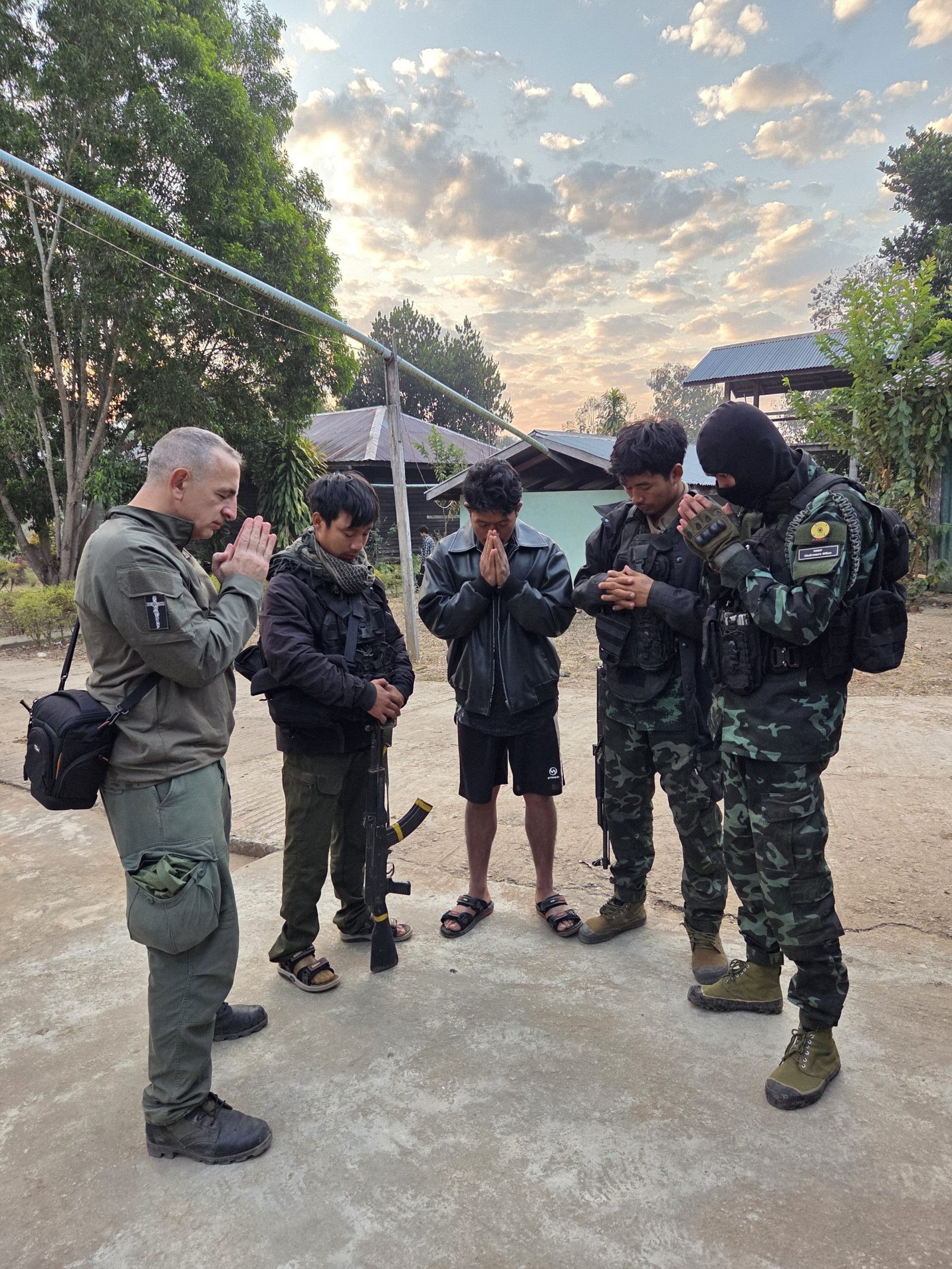 A group of individuals, including armed personnel, bow their heads in prayer during a peaceful moment outdoors at sunrise.