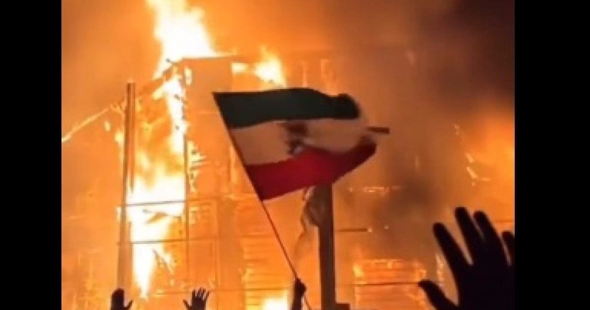Flames engulf a building as a person waves a Mexican flag, symbolizing protest and resilience amidst chaos.