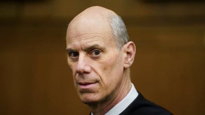 Portrait of a bald man in formal attire, looking directly at the camera with a serious expression, set against a neutral background.