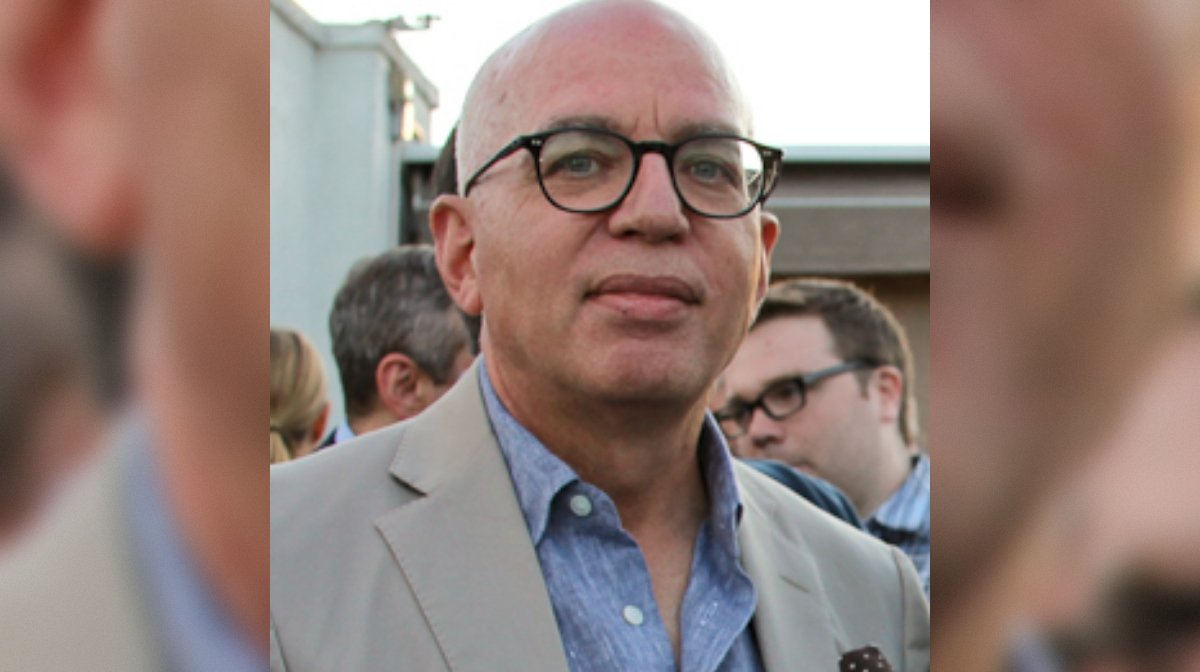 Man wearing glasses and a light-colored suit, standing outdoors among a crowd, with a neutral expression on his face.