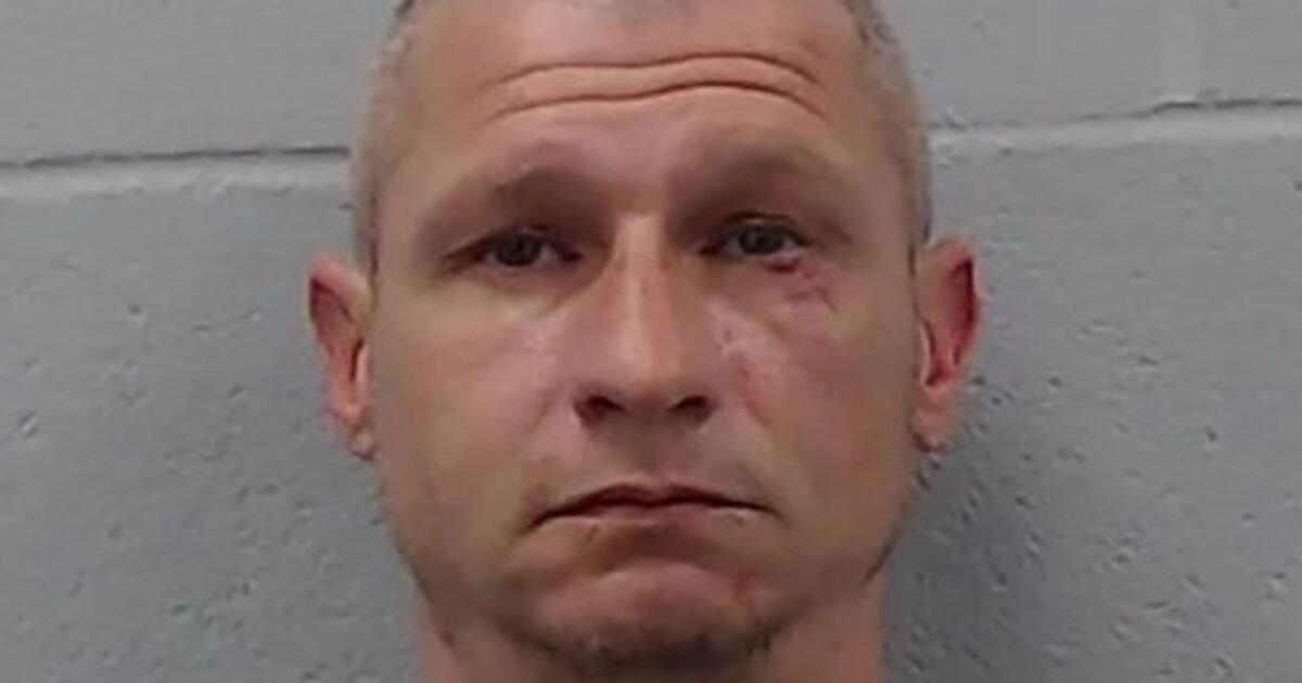 Mugshot of a male individual with short hair and a serious expression, featuring visible facial markings, against a gray background.