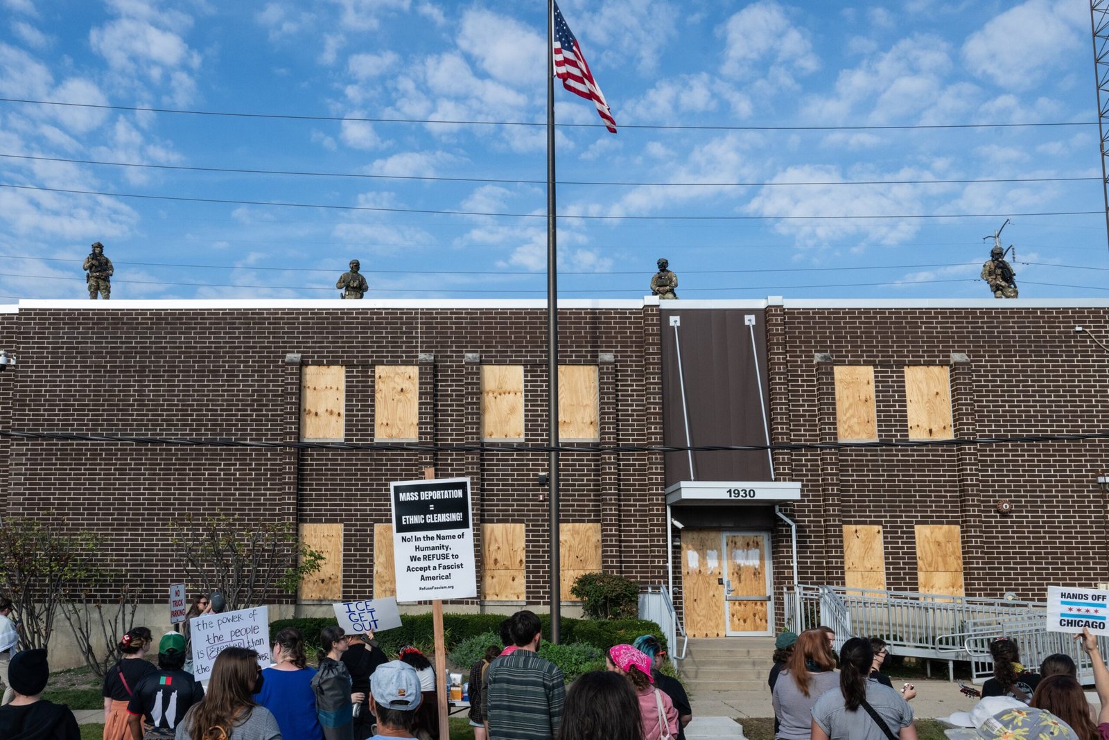 Protesters gather outside a boarded-up building with armed soldiers on the roof, highlighting tensions surrounding immigration policies and community activism.