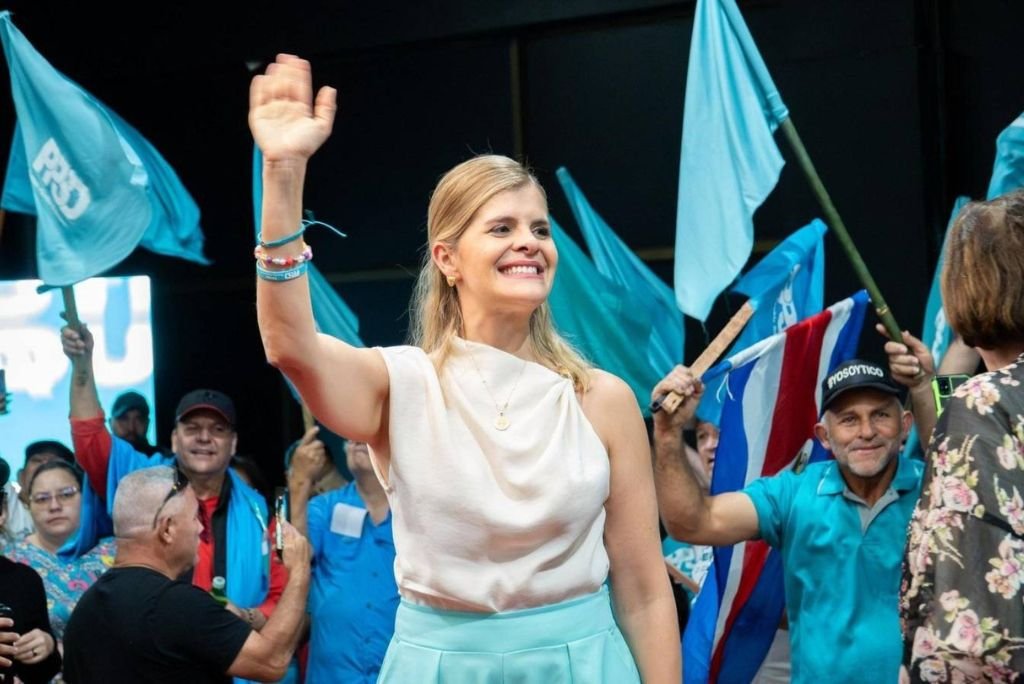 Smiling woman in a white blouse waves to a crowd holding blue flags at a political event, showcasing enthusiasm and support.