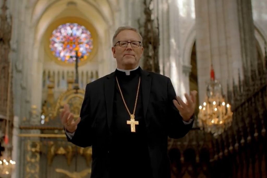 A priest in a black suit and glasses gestures in a grand cathedral with stained glass, symbolizing faith and spirituality.