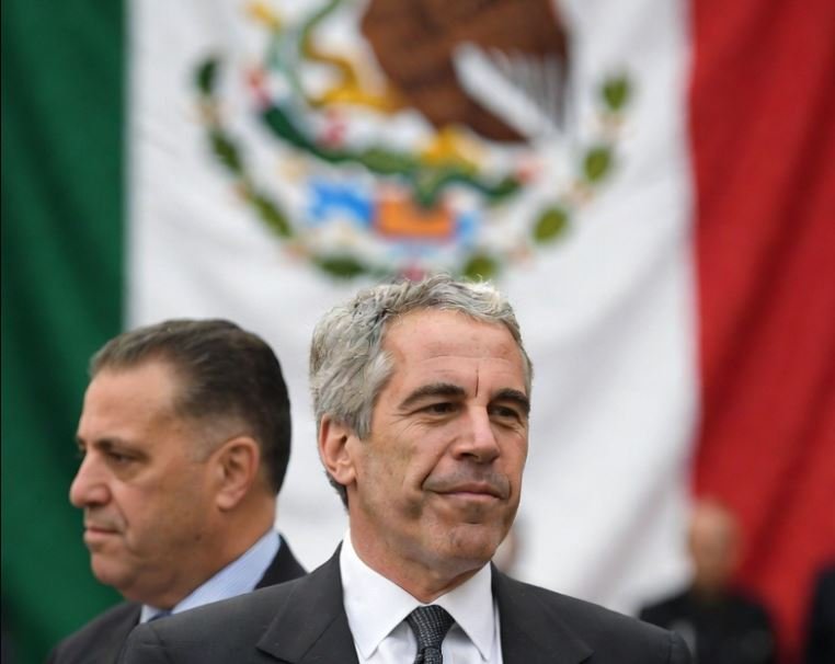 Two men in formal attire stand in front of the Mexican flag, emphasizing a cultural or political event.