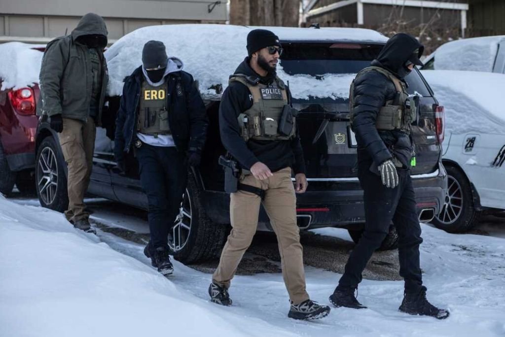 Law enforcement officers in tactical gear walking through snow near parked vehicles during a winter operation.