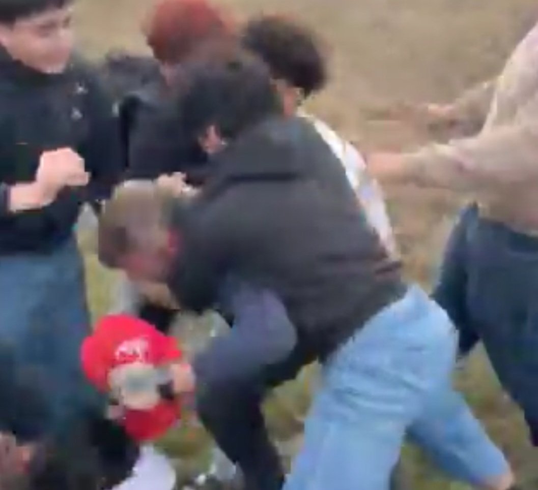 A group of teenagers engage in a physical altercation outdoors, showcasing a chaotic scene with several individuals involved in the struggle.