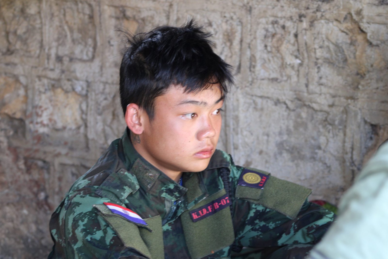 Young soldier in military uniform sitting against a stone wall, displaying a serious expression and wearing insignia patches.