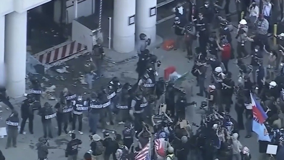 Crowd of protesters clashing with police outside a building, showcasing signs and flags amidst a tense atmosphere.