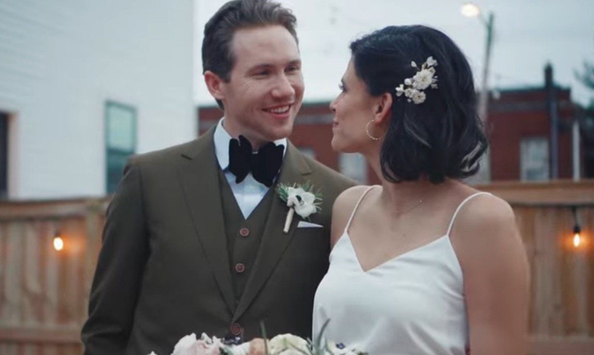 A smiling couple in formal wedding attire stands together outdoors, with the bride holding a bouquet and floral accents in her hair.