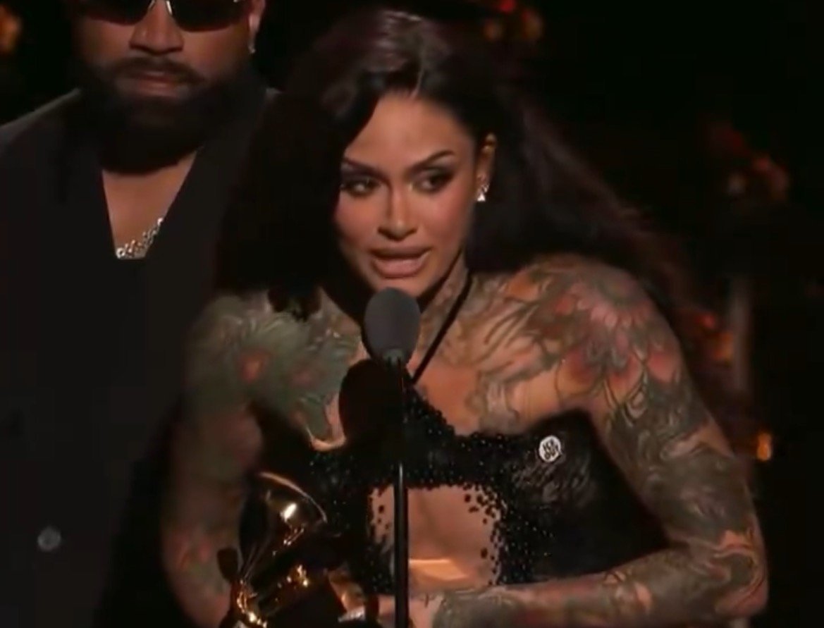 Tattooed artist accepting an award at a ceremony, speaking into a microphone with a male figure in the background.