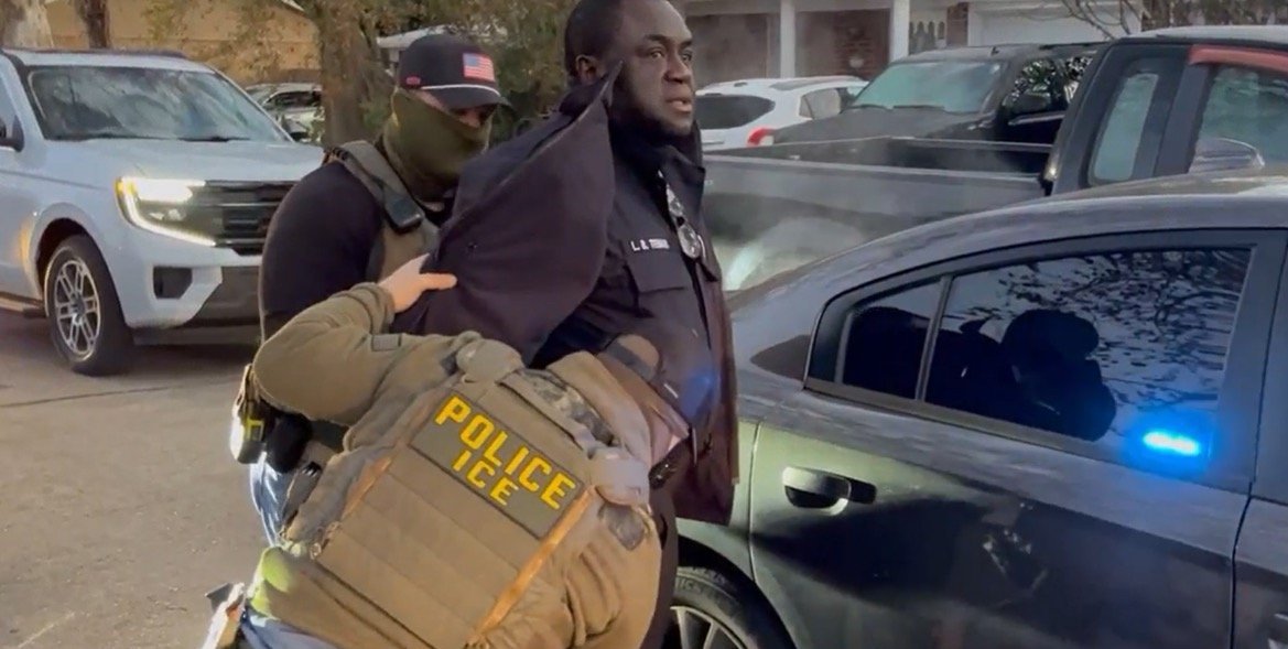 Law enforcement officers apprehend an individual during an operation, with police vehicles and a residential area in the background.