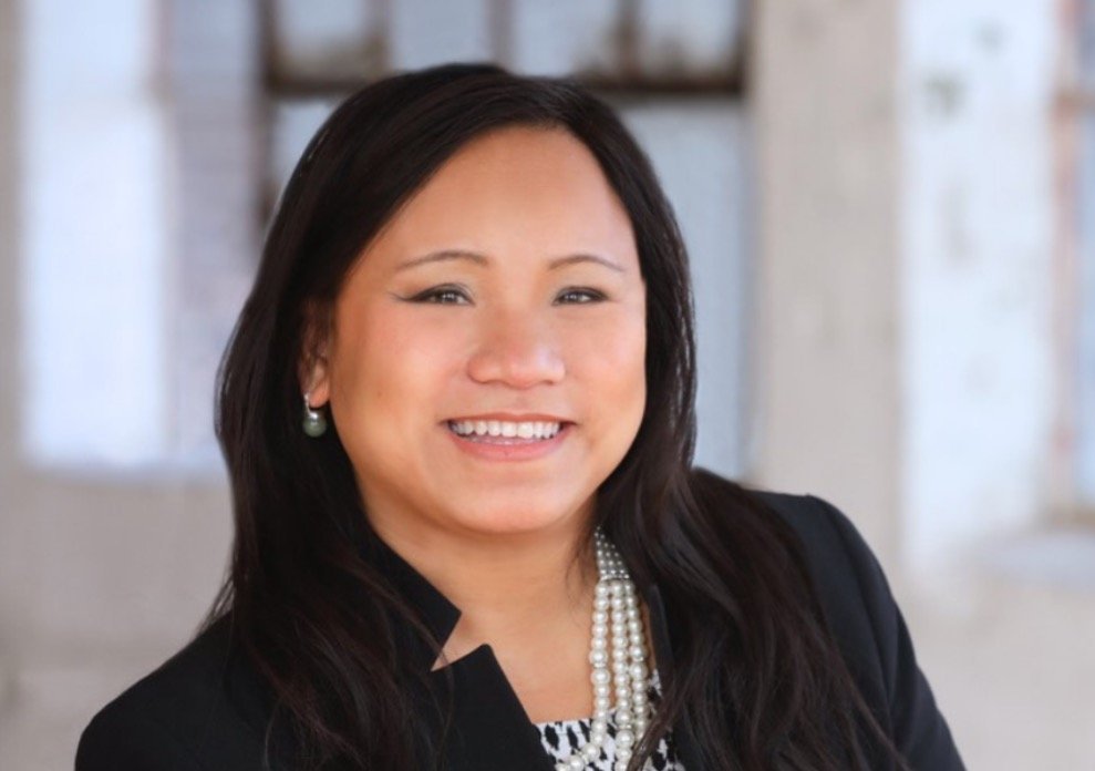 Professional woman smiling in a black blazer and pearl necklace, with a blurred industrial background.