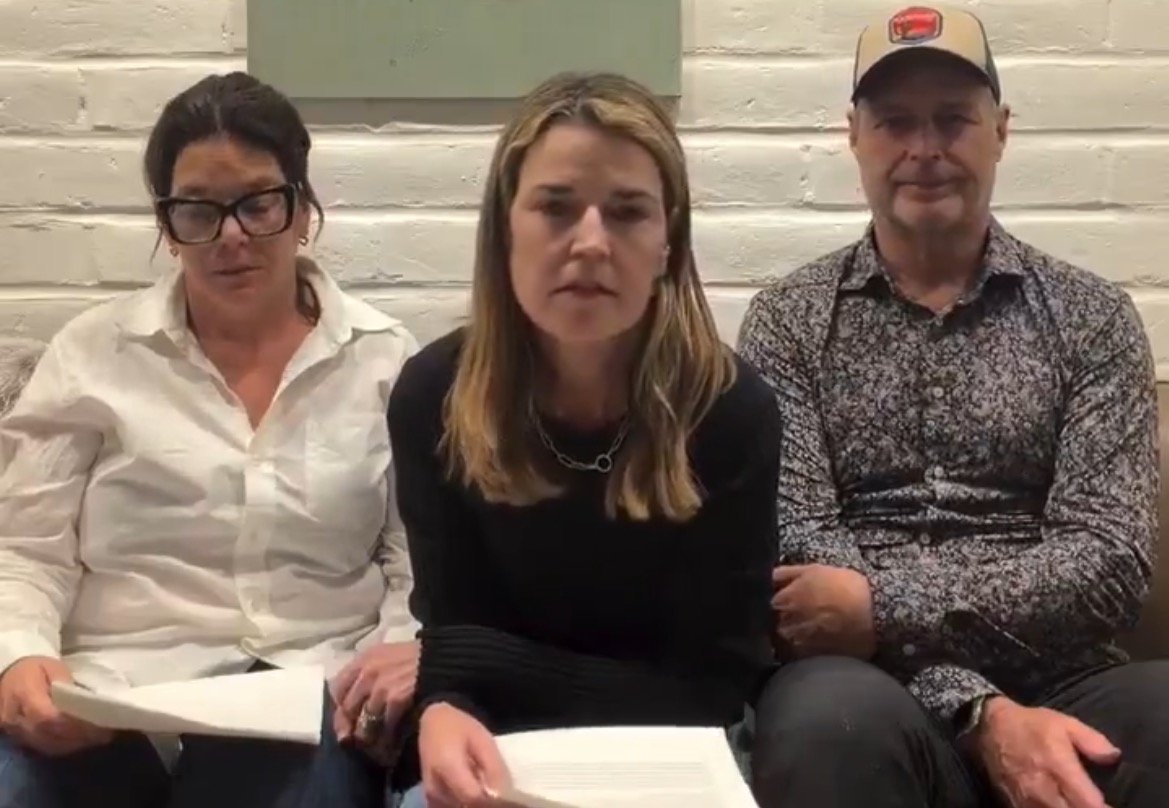 Three individuals seated on a couch, discussing a topic with papers in hand, in a cozy indoor setting with a white brick wall.