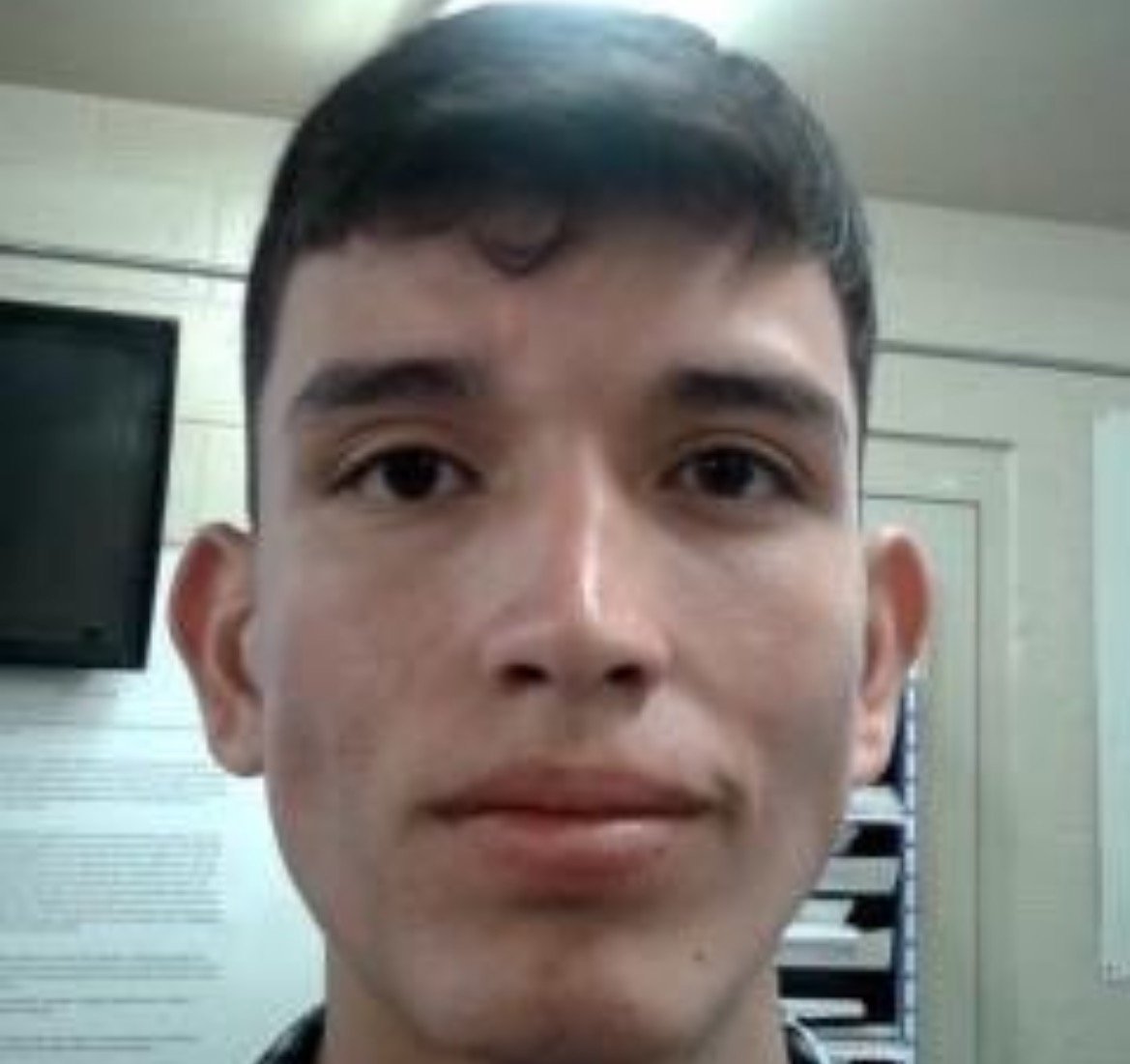 Close-up portrait of a young man with short hair, neutral expression, against a plain background with visible documents and a television screen.