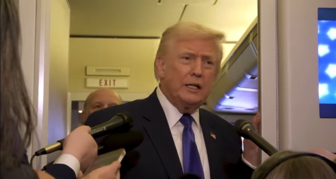 Donald Trump speaking to reporters on an airplane, surrounded by microphones during a press event.