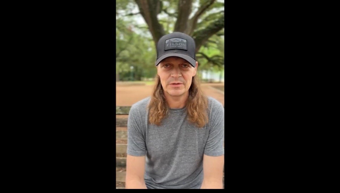 Man with long hair wearing a gray shirt and black cap sits on a bench in a park surrounded by trees.