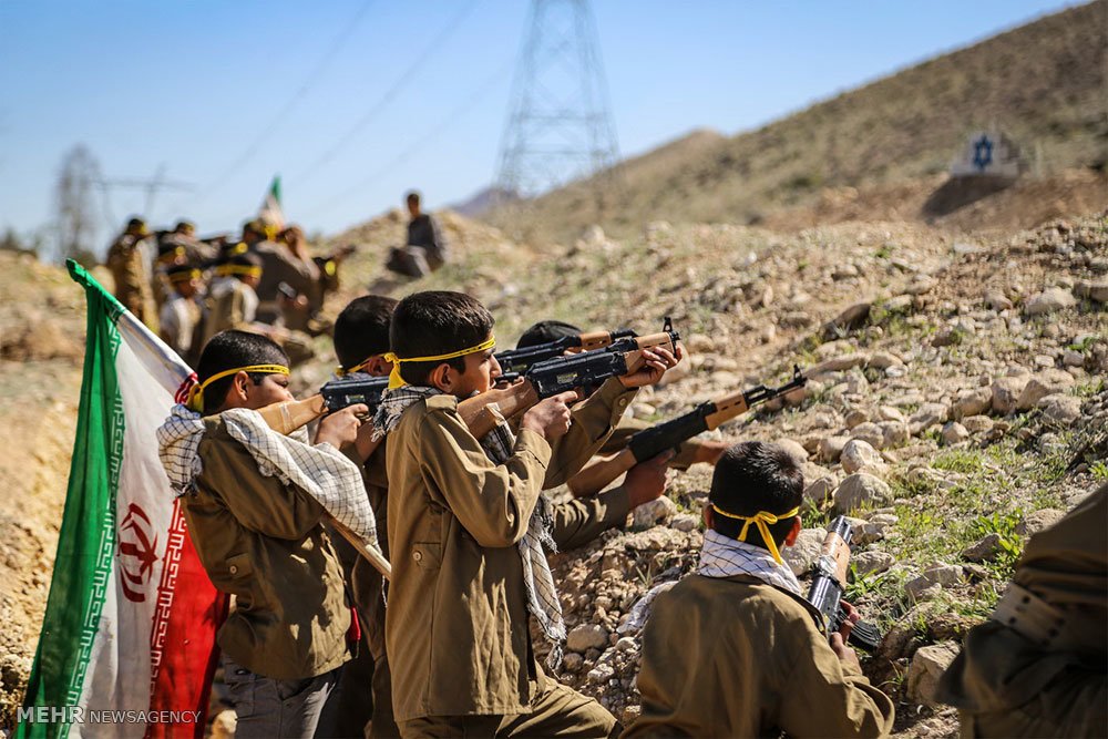 Children in military attire practice shooting with rifles during a training exercise, with flags in the background.