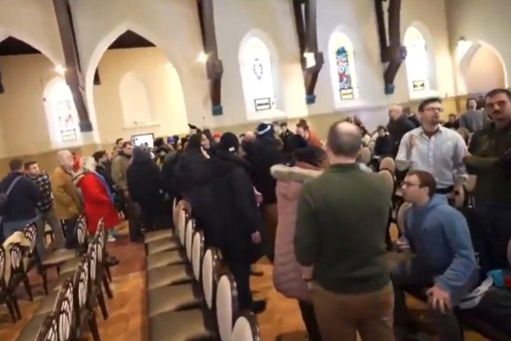 Crowd of people interacting in a spacious hall with wooden beams and stained glass windows, showcasing a lively community event.