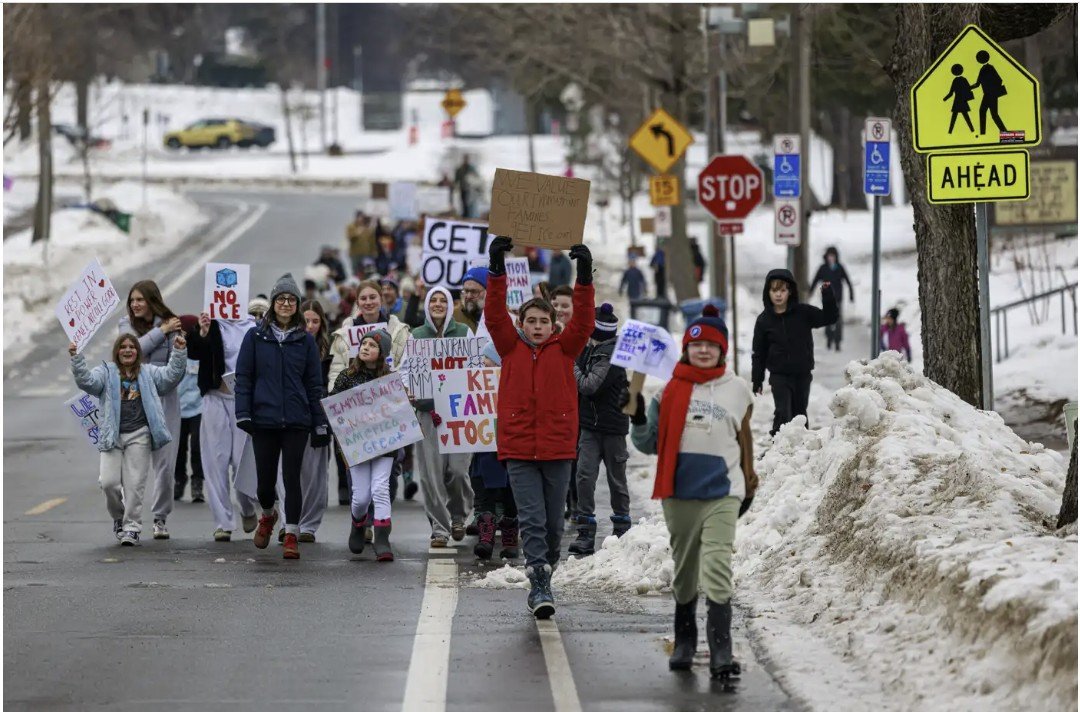 Students and community members march in winter attire holding signs advocating for social change on a snowy street.