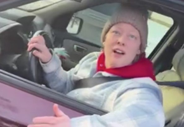 Person in a vehicle wearing a beanie and gray jacket, engaged in conversation while driving, with a visible steering wheel and outdoor background.