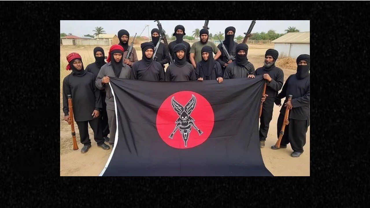 Group of masked individuals in black clothing holding a flag with a distinctive emblem, showcasing armed presence in a rural setting.