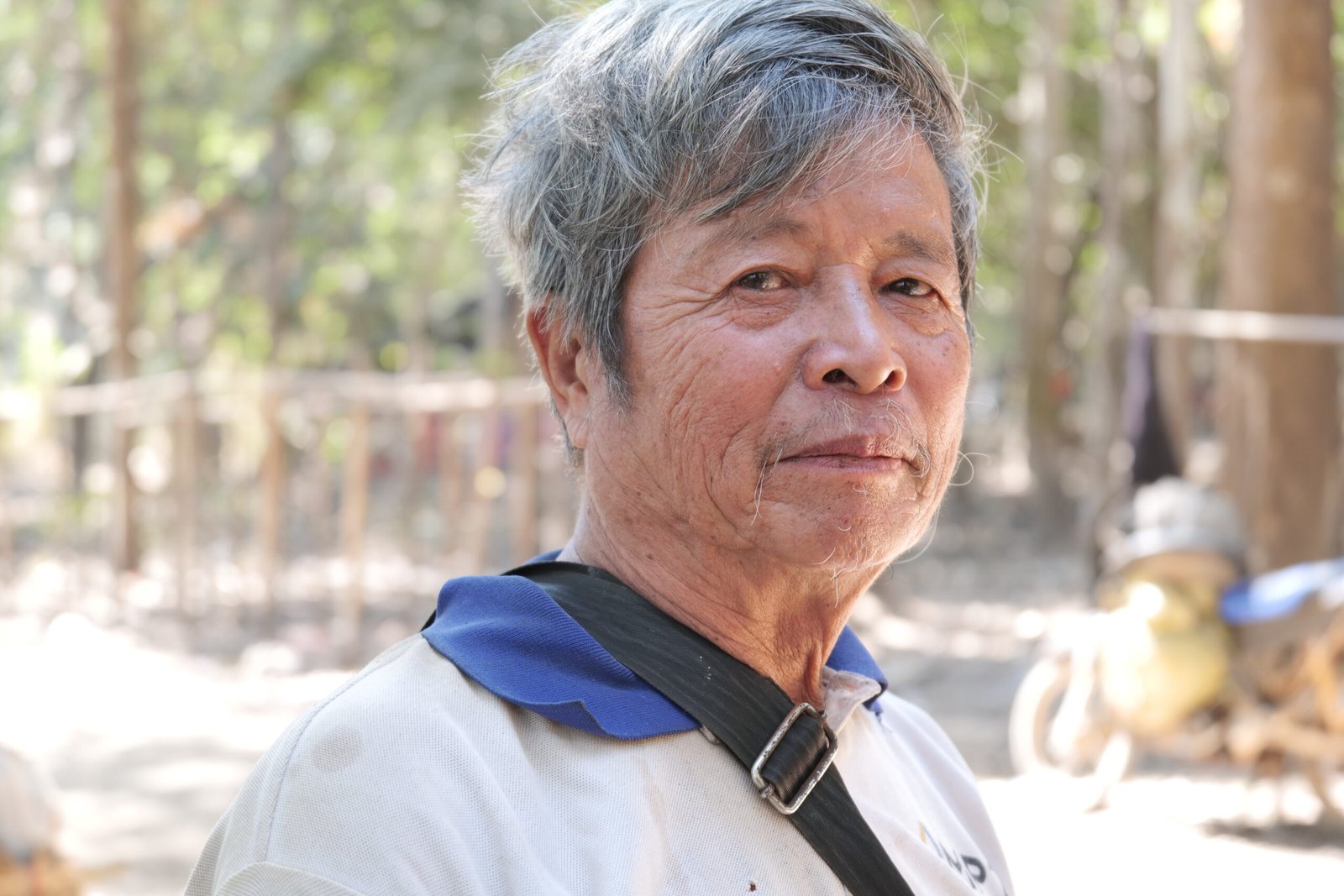 Elderly man with gray hair and a thoughtful expression stands outdoors in a blurred natural setting, wearing a light shirt and black strap.