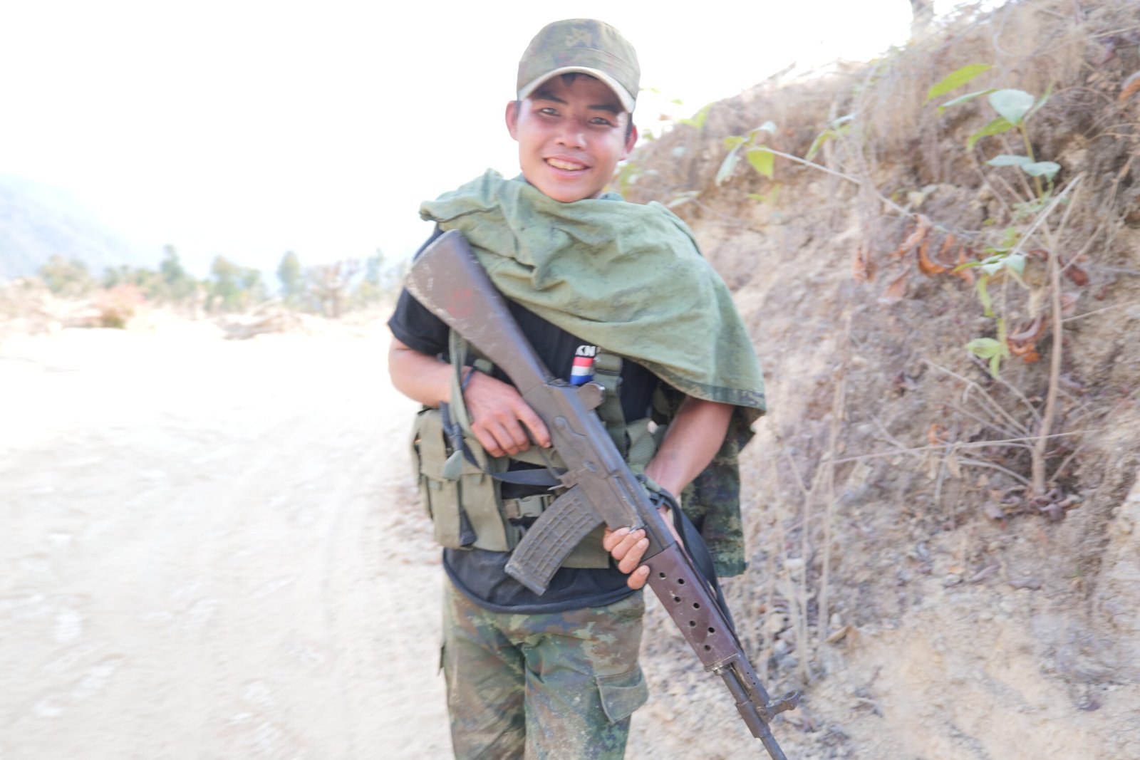 Young soldier in camouflage attire holding a rifle, standing on a dirt path with a mountainous background.