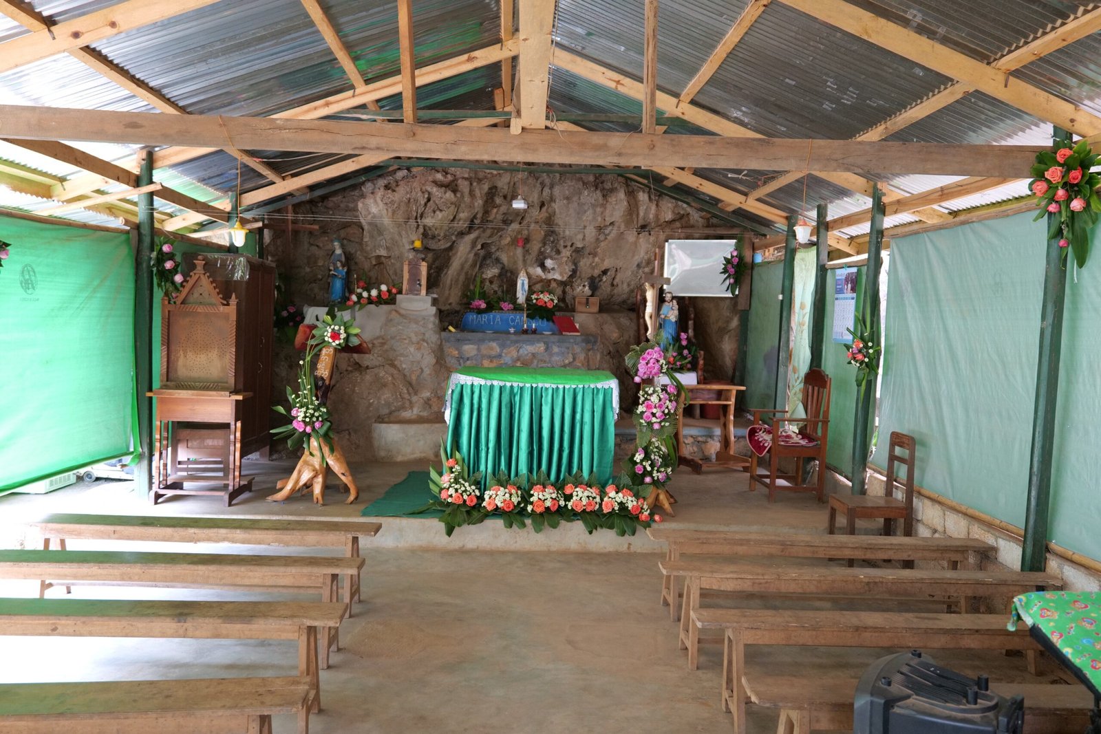 Interior of a modest chapel featuring a rock wall, wooden benches, a decorated altar with flowers, and religious statues, creating a serene atmosphere for worship.