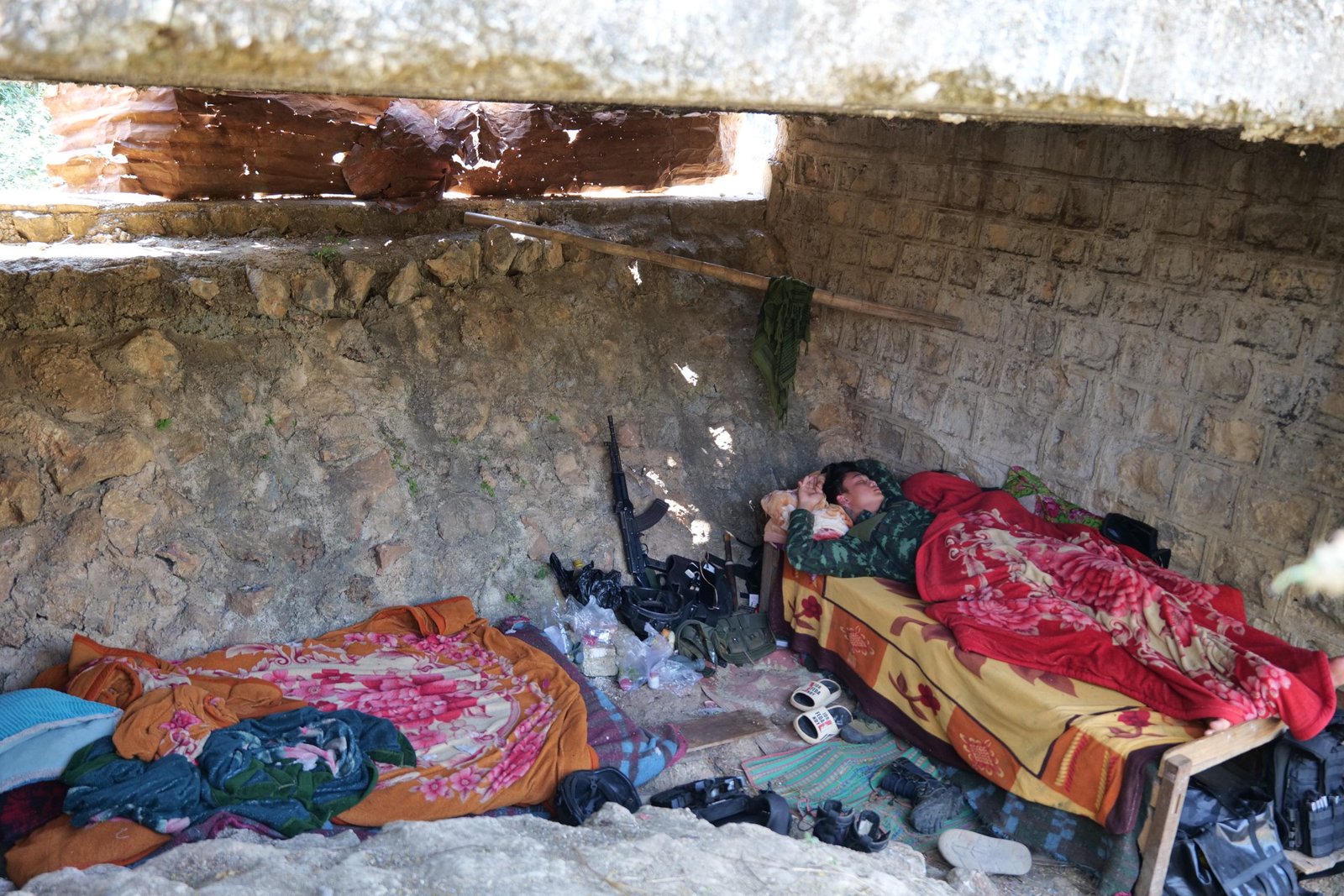 Interior of a makeshift shelter showing two individuals resting on beds surrounded by personal belongings and military gear.