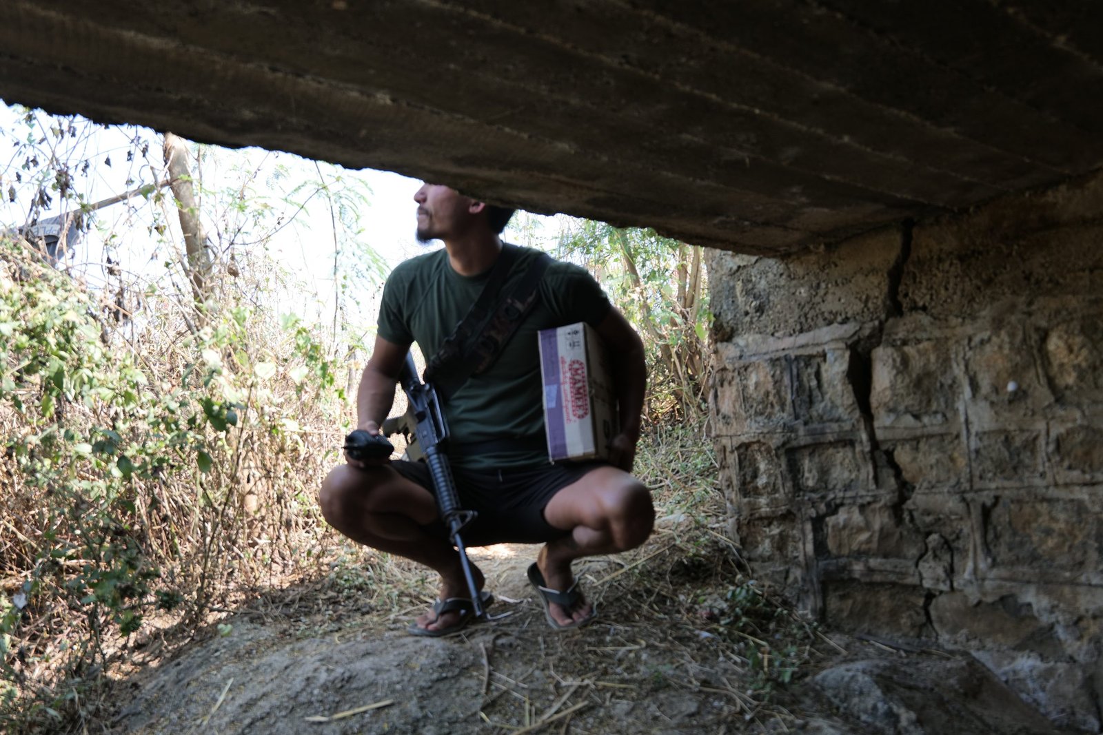 A man crouches under a concrete structure holding a rifle and a box, surrounded by overgrown vegetation.