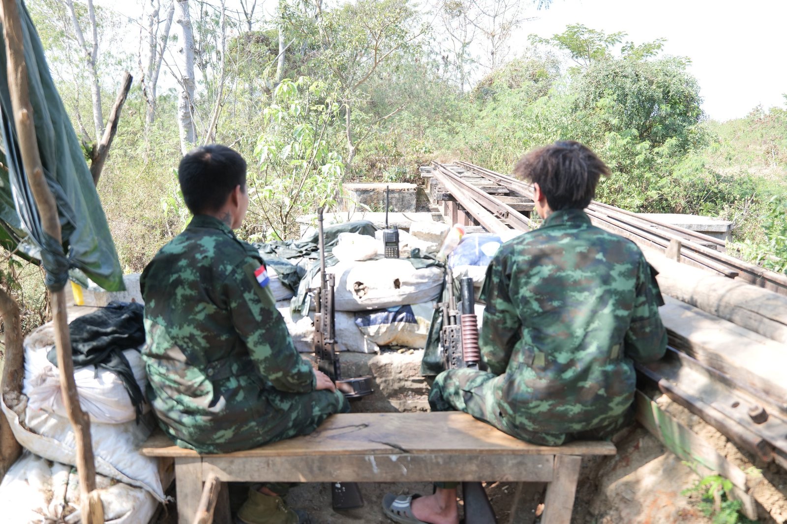 Two soldiers in camouflage uniforms sit on a wooden bench at a lookout point, surrounded by military equipment and greenery.
