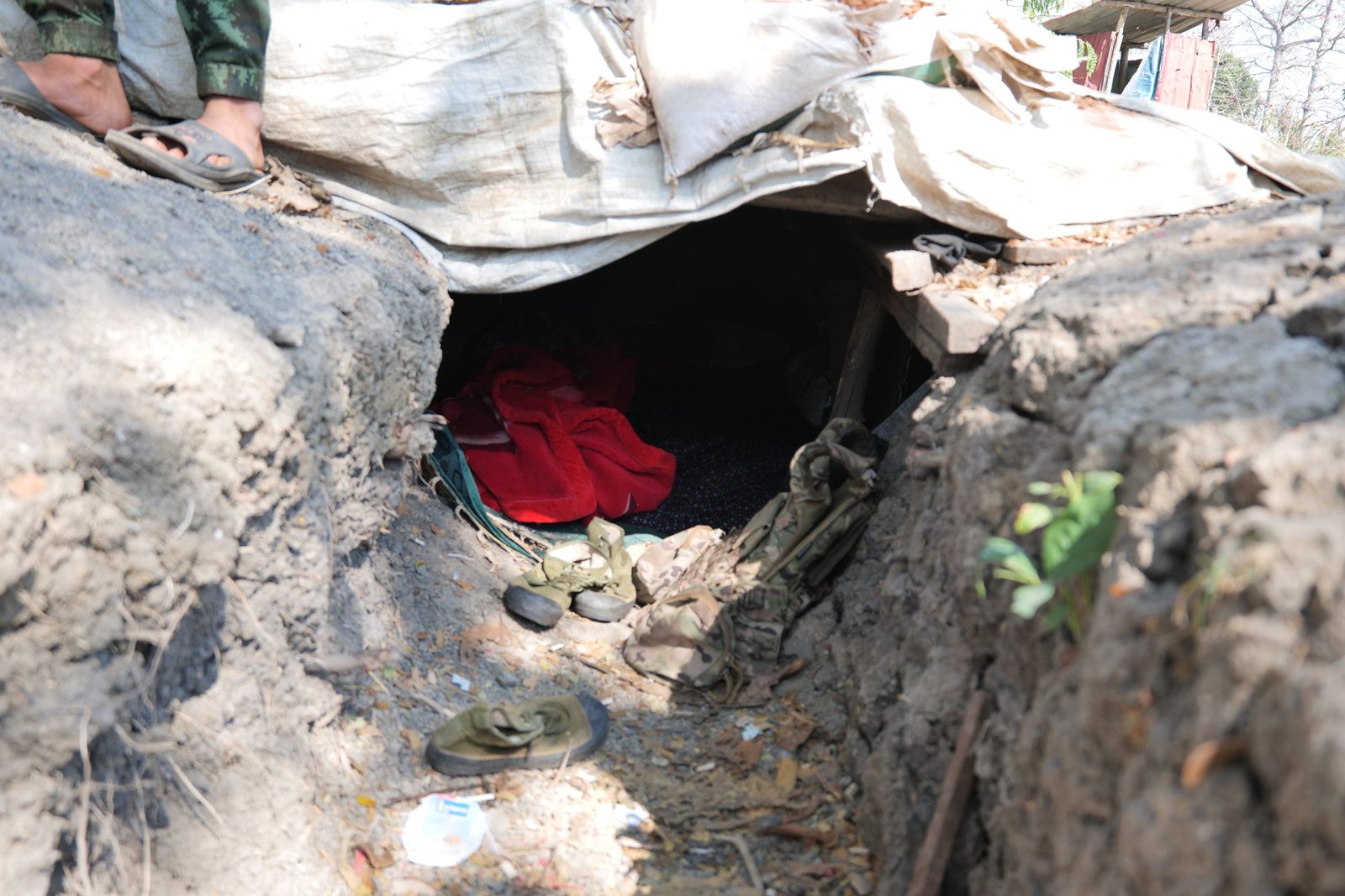 Entrance to a makeshift shelter with red blankets and military gear scattered around, set in a natural environment.