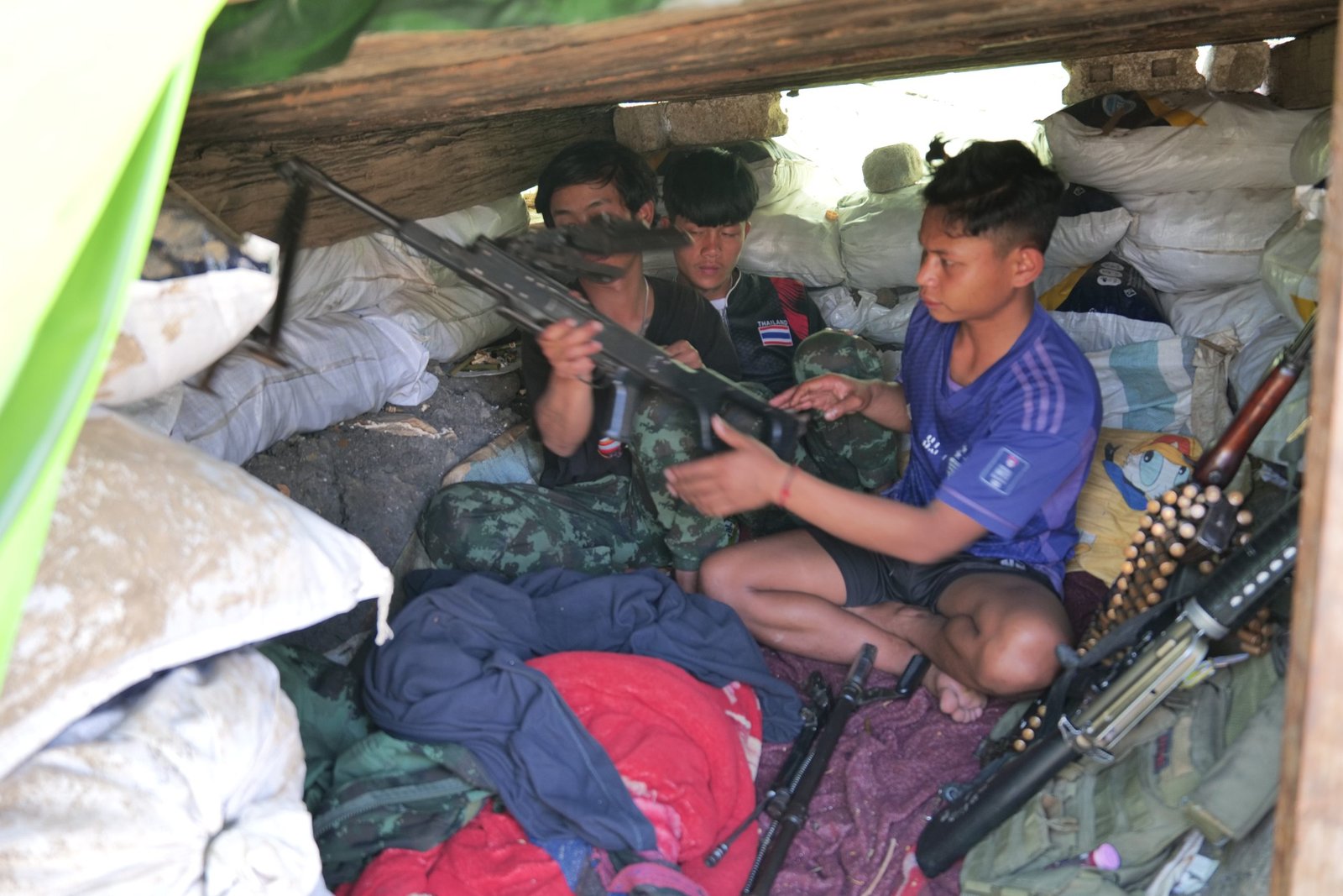 Young individuals in a makeshift shelter handling firearms amidst bags and clothing, highlighting a scene of conflict or military training.