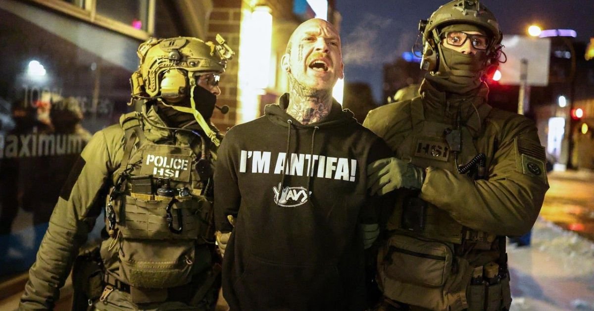 Police officers detain a man wearing an "I'm Antifa!" hoodie during a nighttime protest, highlighting tensions between law enforcement and activist groups.
