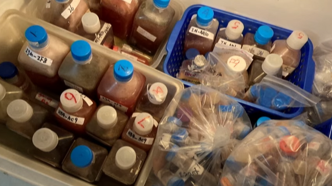 Bottles and bags of labeled chemical samples stored in bins, showcasing organized laboratory materials for research purposes.