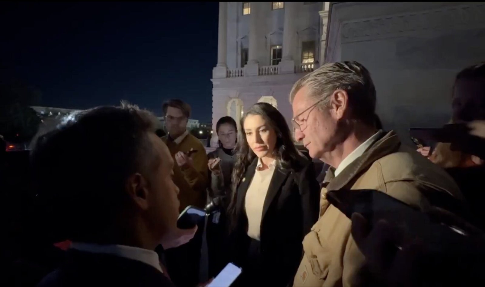 A group of journalists interviews two individuals outside a historic building at night, capturing a moment of discussion amid a media event.